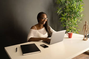 Fröhliche afroamerikanische Freiberuflerin, die an einem Tisch an einem modernen Arbeitsplatz sitzt und auf einem Netbook surft, während sie von zu Hause aus an einem Projekt arbeitet - ADSF29843