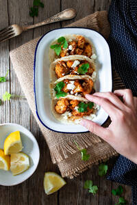 Von oben Hand mit Blumenkohl-Snack-Tacos in Schale und Zitronenstücken in Teller auf Holztisch - ADSF29914