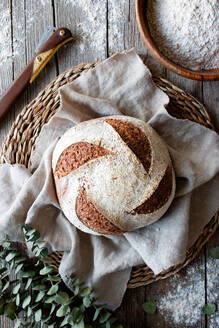 From above homemade fresh sourdough spelt bread on wicker stand with cloth on wooden table with flour scattered - ADSF29923