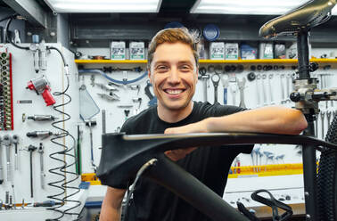 Glücklicher männlicher Mechaniker, der lächelt und in die Kamera schaut, während er sich auf ein zu reparierendes Fahrrad lehnt, das an einer Wand mit Werkzeugen in einer Werkstatt steht - ADSF29933
