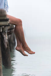 Seitenansicht der Beine einer Frau, die an einem nebligen Morgen an der Playa de Muro auf einem hölzernen Kai am Meer sitzt - ADSF29944