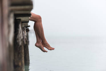 Side view of crop legs of female sitting on wooden quay near sea on misty morning on Playa de Muro - ADSF29945