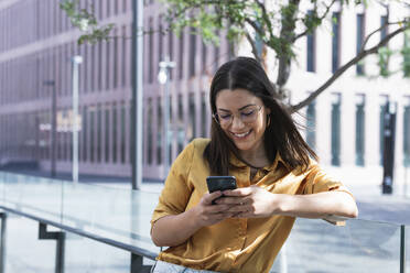 Lächelnde Geschäftsfrau, die ein Mobiltelefon benutzt, während sie sich auf ein Geländer im Büropark stützt - PNAF02156