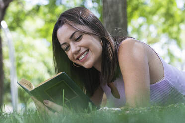 Lächelnde junge Frau liest ein Buch, während sie im Park im Gras liegt - PNAF02162