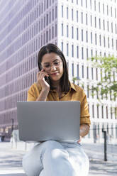 Junge Geschäftsfrau, die mit dem Handy telefoniert und einen Laptop im Büropark benutzt - PNAF02168