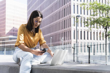 Weibliche Fachkraft, die ein Mobiltelefon benutzt, während sie mit einem Laptop im Büropark sitzt - PNAF02169