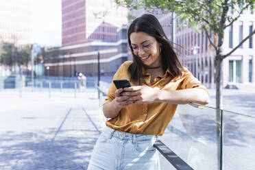 Lächelnde weibliche Fachkraft, die eine Textnachricht über ihr Mobiltelefon sendet, während sie sich auf ein Geländer stützt - PNAF02170