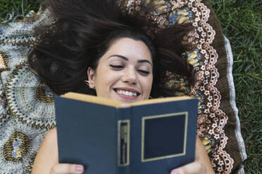 Lächelnde junge Frau liest ein Buch, während sie auf einer Decke im Park liegt - PNAF02189
