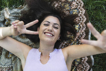 Happy young woman gesturing peace sign while lying at public park - PNAF02190