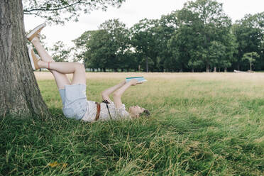 Woman reading book while relaxing on grass at park - ASGF01382
