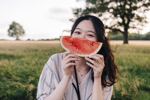 Junge Frau bedeckt ihr Gesicht mit einer Wassermelonenscheibe auf einer Wiese - ASGF01404