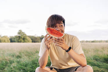 Junger Mann hält Wassermelonenscheibe auf einer Wiese sitzend - ASGF01407