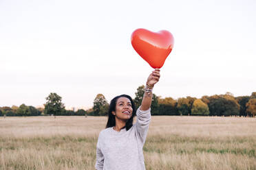 Lächelnde Frau, die einen Ballon in Herzform hält, während sie im Park steht - ASGF01443