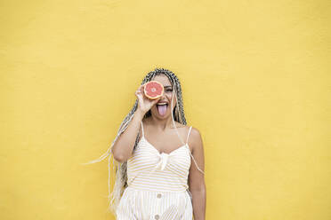 Frau streckt die Zunge heraus, während sie eine Zitrusfrucht vor dem Auge an einer gelben Wand hält - JCCMF03850