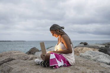 Lächelnde junge Frau mit Laptop auf einem Felsen am Strand bei Sonnenuntergang - JCCMF03858