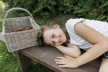 Lächelndes Mädchen neben Weidenkorb auf Bank im Garten liegend - LLUF00003