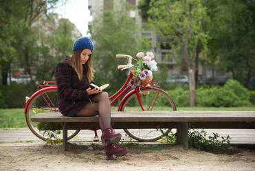 Eine Frau liest ein Buch, ein rotes Fahrrad und ein Korb mit bunten Blumen. - CAVF94642