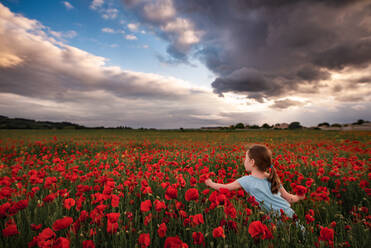 Mädchen pflückt Mohnblumen auf einem Feld mit Sonnenuntergangswolken am Himmel in Frankreich - CAVF94651