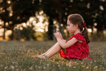 Girl sits on grassy lawn examining clover flower in evening sun - CAVF94653