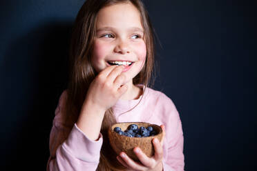 Lächelndes Mädchen pflückt frische Blaubeeren auf dunklem Hintergrund - CAVF94662