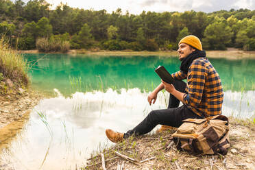 Spanish traveler enjoying reading a book sitting on a peaceful lake - CAVF94712