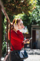 Young red-haired woman relaxing while listening to music with her cell phone and yellow headphones in an urban space. Dressed in a red blouse. - CAVF94723