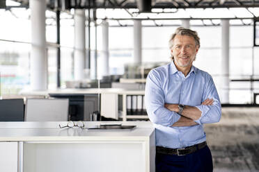 Smiling male professional with arms crossed leaning on desk at office - PESF03165