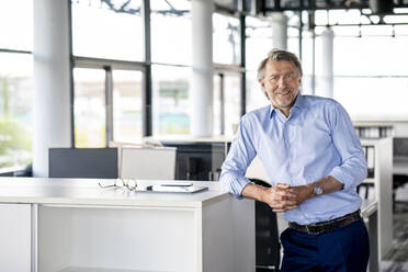 Smiling male professional with hands clasped leaning on desk - PESF03166