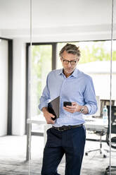 Businessman wearing eyeglasses using mobile phone at office - PESF03168