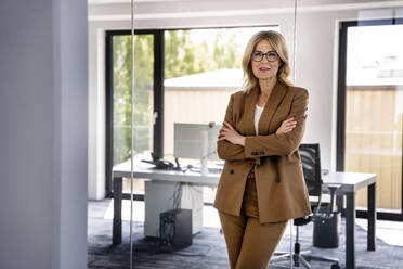 Confident businesswoman with arms crossed leaning on glass in office - PESF03179