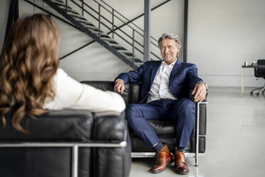 Lächelnder Geschäftsmann, der eine Kollegin ansieht, während er auf dem Sofa im Büro sitzt - PESF03208