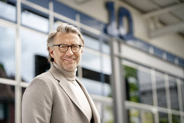 Älterer Geschäftsmann mit Brille steht vor einem Gebäude im Büropark - PESF03218