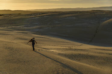 Männlicher Tourist, der ein Stativ trägt, während er bei Sonnenuntergang über den Sand läuft - TOVF00278
