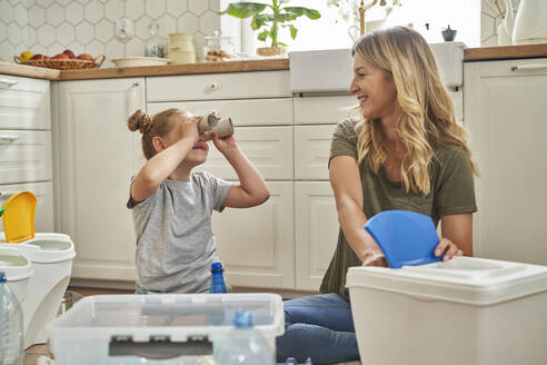 Playful girl looking at mother through rolled up paper in kitchen - ABIF01592