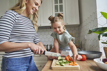 Smiling mother cutting vegetables by daughter sitting on kitchen counter - ABIF01602