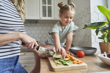 Girl taking chopped cucumber while mother preparing food in kitchen - ABIF01603