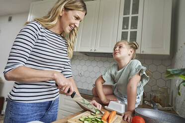 Smiling mother looking at daughter while chopping cucumber in kitchen - ABIF01604