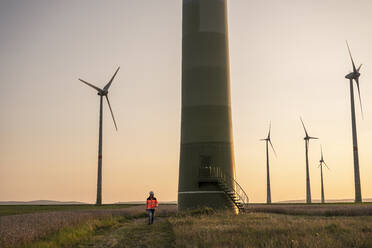 Male engineer walking on meadow in front of wind turbines - UUF24604