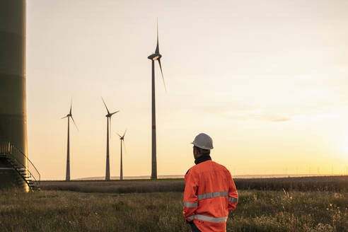 Männlicher Inspektor betrachtet Windkraftanlagen auf einer Wiese bei Sonnenuntergang - UUF24606