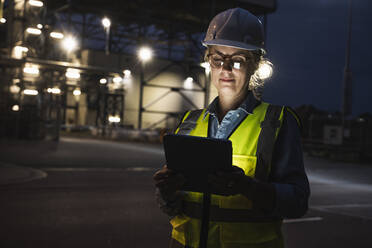 Female engineer with hardhat using digital tablet outside factory at night - UUF24608
