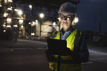 Ingenieurin mit Schutzhelm bei der Arbeit mit einem digitalen Tablet außerhalb einer Fabrik bei Nacht - UUF24608