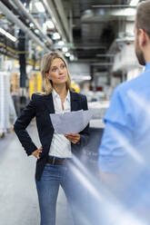 Weiblicher Geschäftsmann mit Hand auf der Hüfte, der einen Kollegen in einer Fabrik betrachtet - DIGF16493