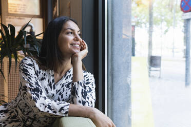 Lächelnde junge Frau träumt, während sie durch das Fenster eines Cafés schaut - PNAF02208