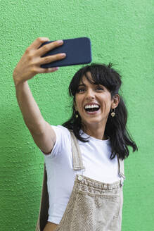Fröhliche Frau nimmt Selfie durch Smartphone gegen grüne Wand - PNAF02231