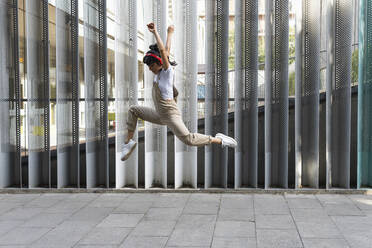 Excited young woman with arms raised jumping on footpath - PNAF02241