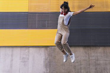 Cheerful woman with arms raised jumping in front of wall - PNAF02243