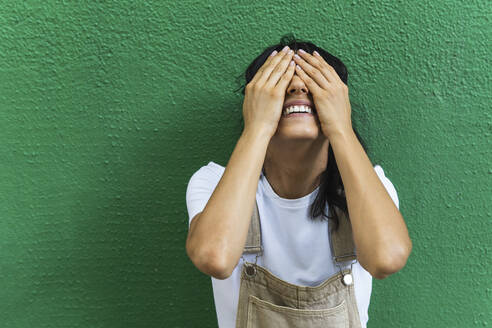 Lächelnde junge Frau, die ihre Augen mit den Händen vor einer grünen Wand bedeckt - PNAF02245