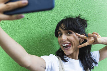 Fröhliche Frau zeigt Friedenszeichen und streckt die Zunge heraus, während sie ein Selfie macht - PNAF02247