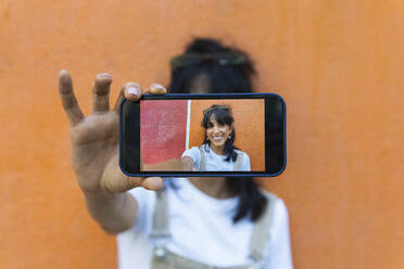 Junge Frau zeigt ein Foto auf ihrem Smartphone vor einer orangefarbenen Wand - PNAF02253