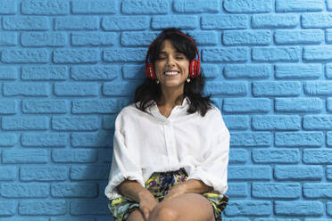 Lächelnde Frau mit geschlossenen Augen hört Musik vor einer blauen Backsteinmauer - PNAF02255